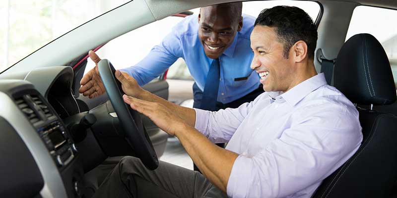 Two men in a car smile and point at the steering wheel, enjoying their time together.